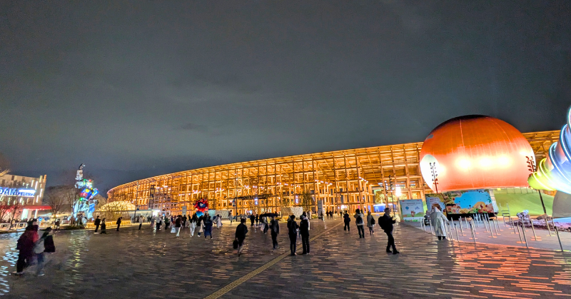 大阪万博会場に設置された「200mの大屋根リング」の夜景。ライトアップされた木組みの大屋根と、来場者が歩く広場、右手に見える赤い球体パビリオンの景観
