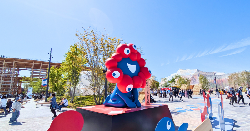 晴天の下、多くの人でにぎわう大阪万博会場に展示された公式キャラクター「ミャクミャク」のカラフルな立体オブジェ