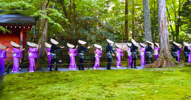 「京都・三千院の境内で行われる伝統行事。紫色の着物と編笠を身に着けた女性たちが整列し、緑豊かな苔庭の中を静かに進んでいる様子。」