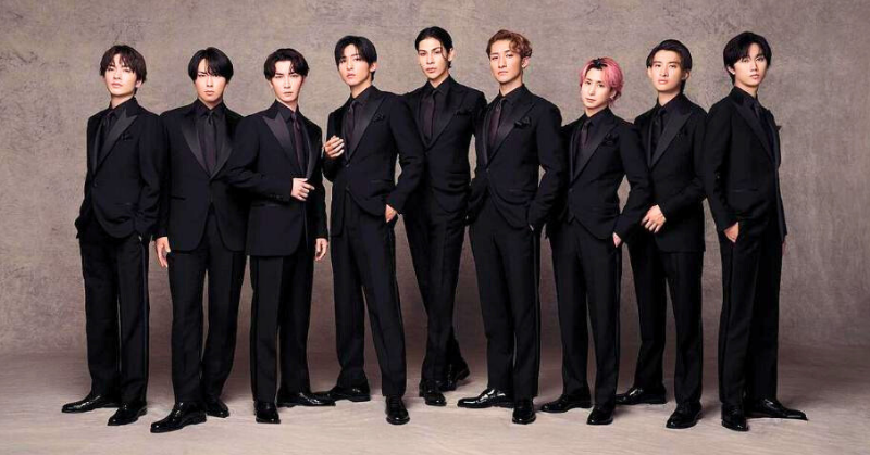 黒いスーツを着た9人の男性アイドルグループsnowmanが横一列に並んで立っている。背景はシンプルなグレーのスタジオ背景で、全員がフォーマルかつスタイリッシュな印象。