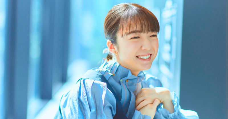 明るい表情で笑顔を見せる。上白石萌音。青いシャツを着て、自然光の差し込む室内でリラックスした様子