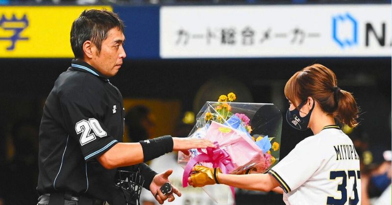 「プロ野球の試合前セレモニーで、審判に花束を手渡すユニフォーム姿の女性スタッフ。審判は真剣な表情で花束を受け取っている。」