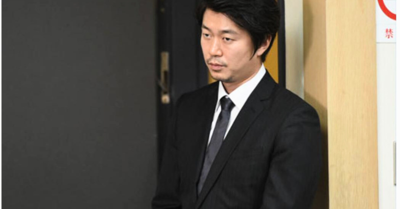 黒いスーツを着た新井浩文が、無表情で壁際に立っている画像