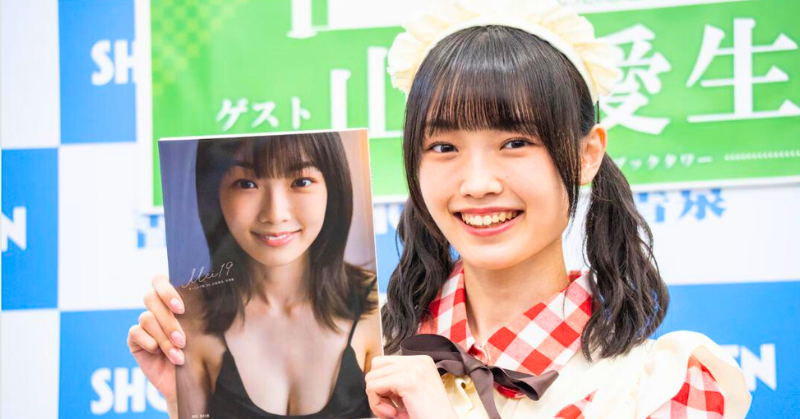 メイド風の衣装を着た山崎愛生が、笑顔で自身の写真集を手に持って紹介している様子