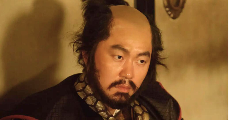 時代劇の衣装を着た新井浩文が険しい表情で座っている。額の広い髪型（ちょんまげ）と髭が特徴的な画像