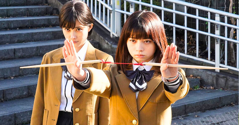 「制服姿のが桜田ひよりが割り箸を両手で強く握り、真剣な表情で構えている。後ろにはもう一人の女子学生が見守っている。」