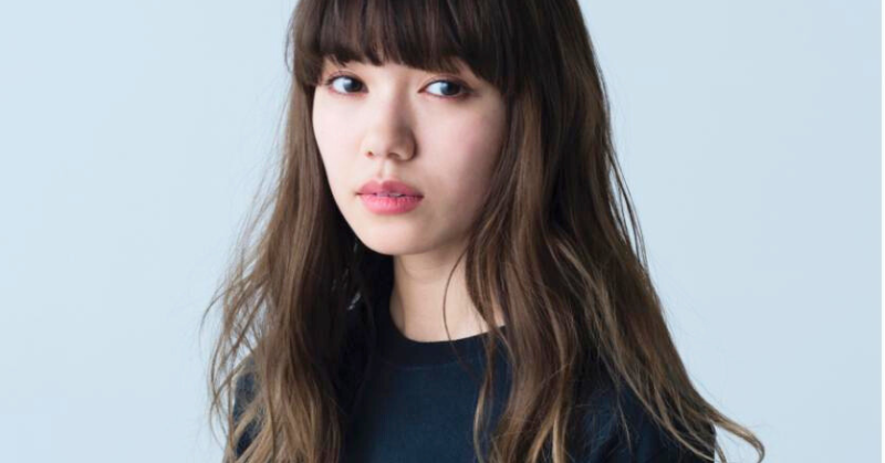 長い茶髪と前髪が特徴的な二階堂ふみが、無表情でカメラを見つめている。シンプルな黒い服を着て、淡い水色の背景の前に立っている画像