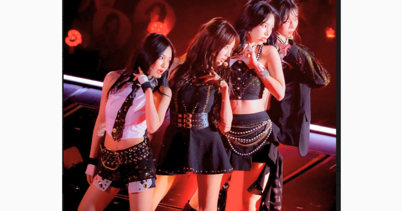 aespaの4人のメンバーが、赤いライトに照らされたステージ上でパフォーマンス中。全員がブラックを基調にした衣装を着用し、真剣な表情で指を口元に当てる印象的なポーズをとっている画像