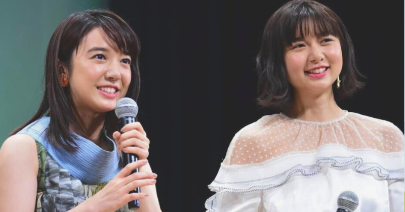 2人の若い女性がマイクを持って笑顔で話している様子。左の萌音はノースリーブのカラフルなドレスを着用し、右の萌歌は白いフリルとドット柄のブラウスを着ている。背景は暗く、ステージ上のイベントかインタビューの場面