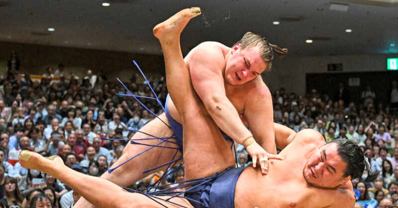 大相撲の激しい取組で、安青錦関（上）が相手力士を投げ技で倒す瞬間。体ごと空中に舞い上がり、観客が固唾をのんで見守っている。