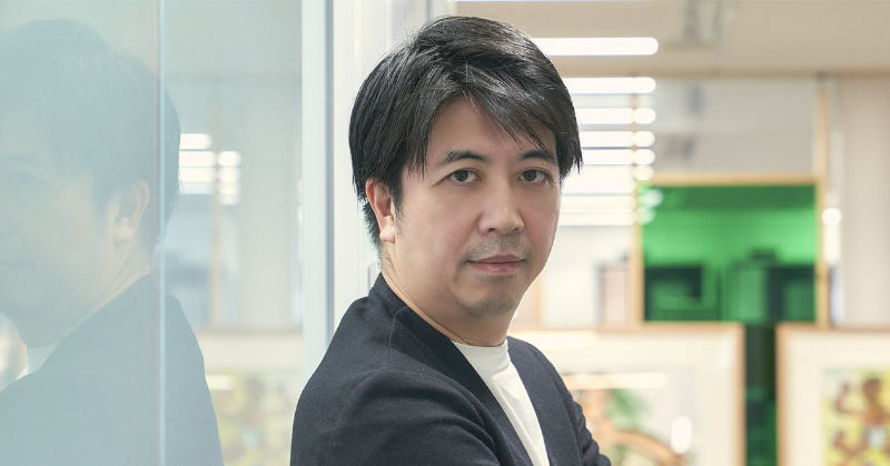 オフィスのガラス壁の前で腕を組み、カメラ目線で立つ黒髪の男性のポートレート。